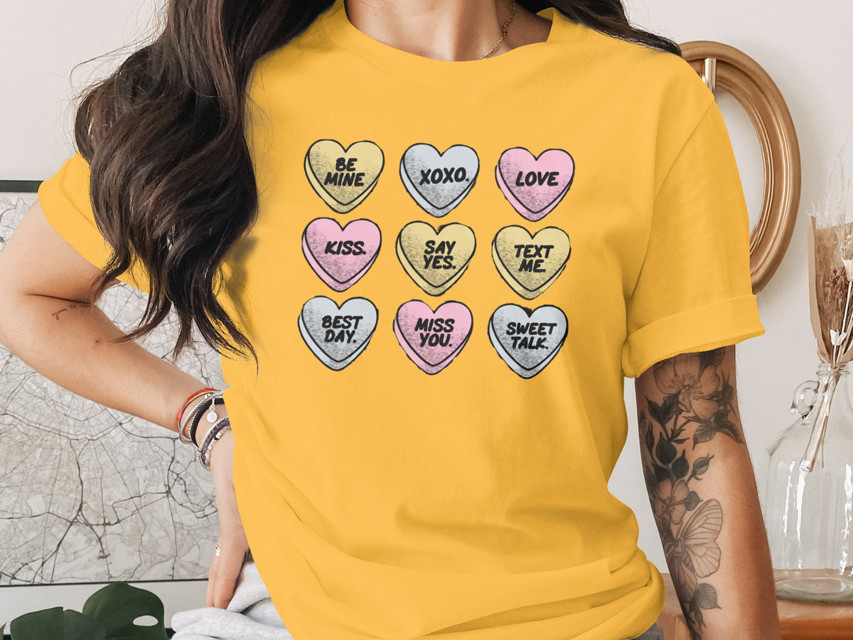 A woman wears a yellow t-shirt printed with a grid of pastel conversation hearts bearing romantic phrases.