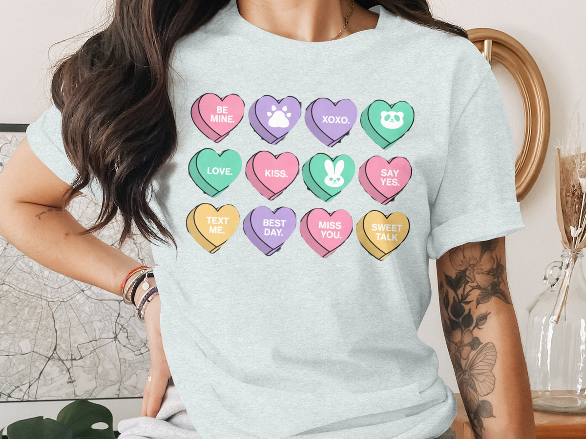 A light blue t-shirt featuring twelve colorful conversation heart candies printed across the chest with various text and icons.