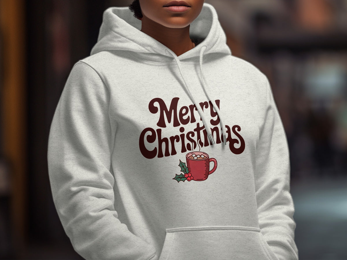 A person wears a white hooded sweatshirt printed with brown retro-style text reading Merry Christmas and a steaming red mug.