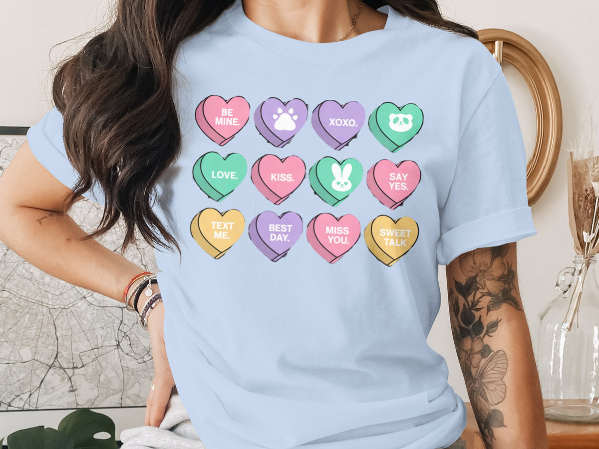 A light blue t-shirt featuring twelve colorful conversation heart candies printed with various Valentine-themed messages and icons.