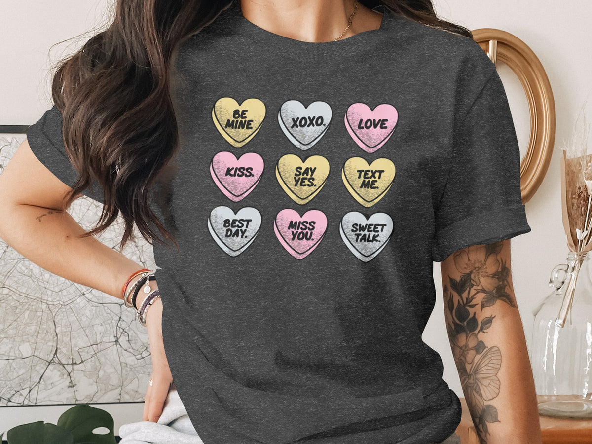 A person wears a dark gray t-shirt printed with a grid of pastel conversation hearts featuring phrases like be mine and love.