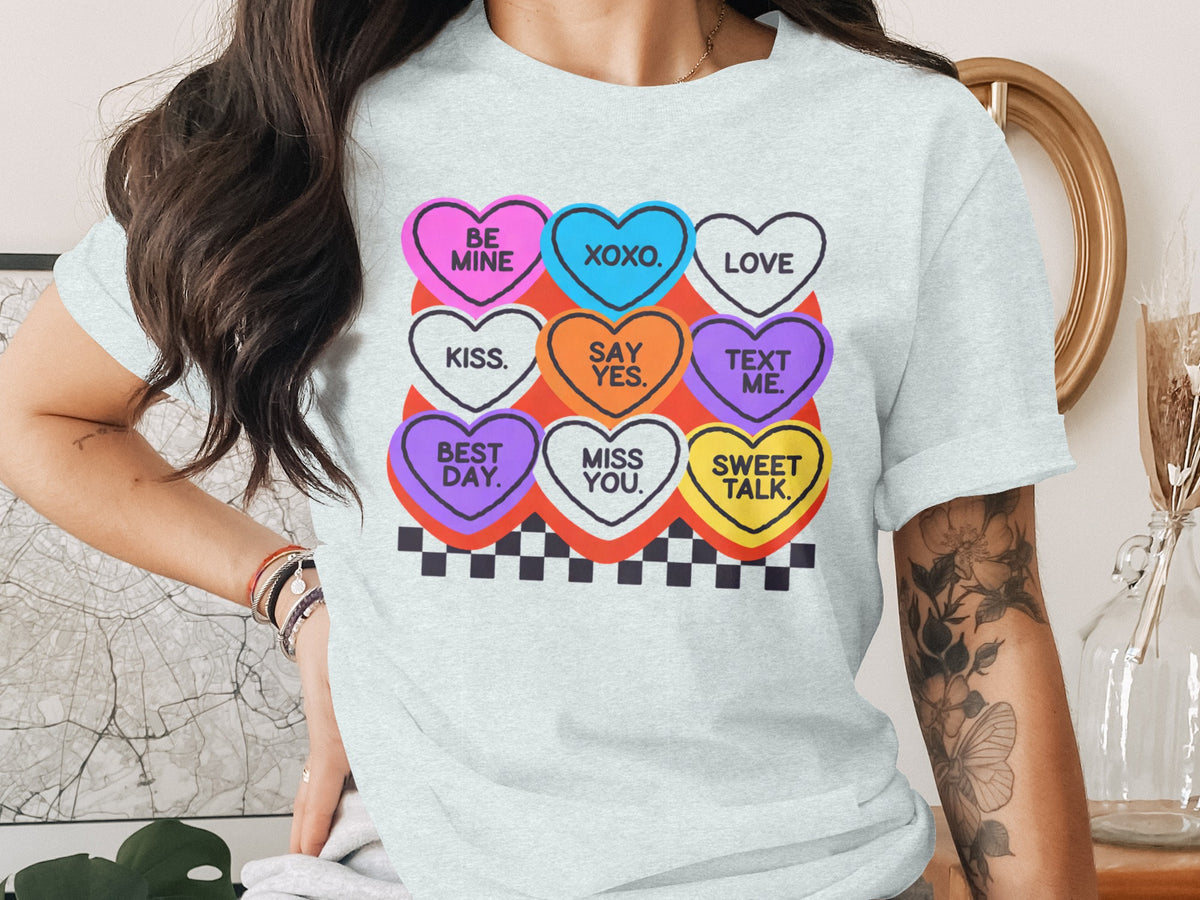 A person wears a light gray t-shirt featuring a grid of colorful candy hearts with messages.