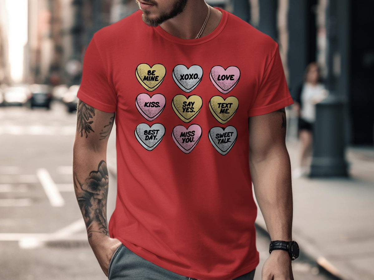 A man wearing a red t-shirt printed with a 3x3 grid of colorful candy hearts featuring phrases like "Be Mine" and "XOXO".