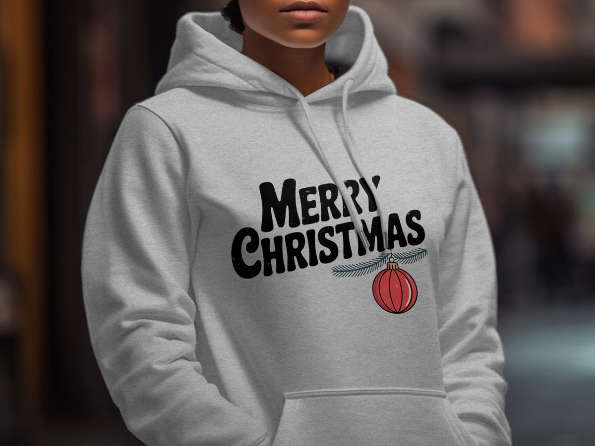 A person wearing a light gray hooded sweatshirt with black text reading MERRY CHRISTMAS and a hanging red ornament graphic.