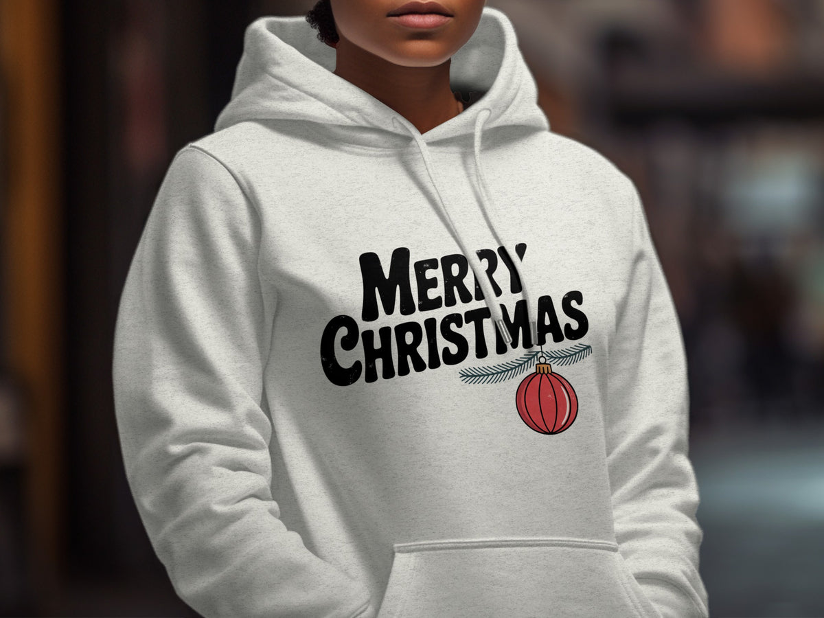 A person wearing a light gray hoodie printed with black text reading MERRY CHRISTMAS and a hanging red ornament.